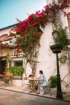 two people sitting at a table in front of a building with flowers growing on it