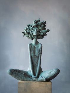 a statue sitting on top of a wooden block in front of a gray wall with a plant growing out of it's head