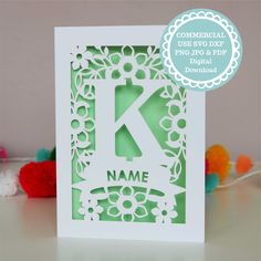 a white card with the letter k cut out in it's center and surrounded by pom poms