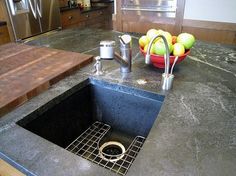 a kitchen sink sitting under a counter top next to a cutting board and bowl of fruit