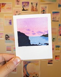 a hand holding up a small card with watercolors on the paper and pictures behind it