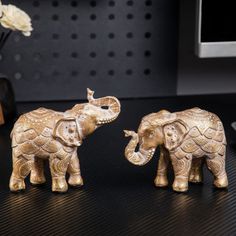 two small gold elephants standing next to each other on a black table with flowers in the background