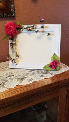 a white frame with pink flowers and green leaves on top of a wooden table next to a brown wall