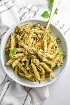 a white bowl filled with pesto pasta on top of a striped cloth next to a wooden spoon