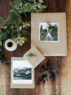 two wrapped gifts sitting on top of a wooden table next to greenery and a roll of tape