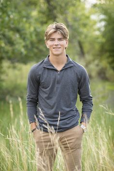 a young man standing in tall grass with his hands in his pockets and smiling at the camera