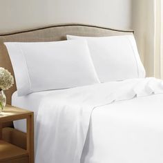 a bed with white sheets and pillows on top of it next to a nightstand in front of a window