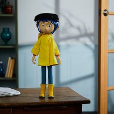 the doll is wearing a yellow raincoat and blue jeans, standing on a wooden table