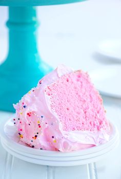 a slice of pink cake with sprinkles sits on a plate next to a blue cake stand