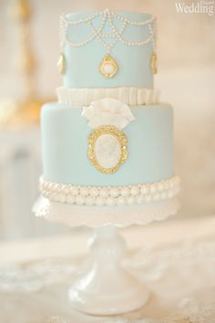 a three tiered blue cake with pearls on the bottom and gold trimmings