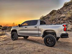 a silver truck parked on top of a dirt road next to a mountain at sunset