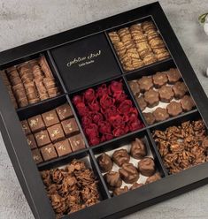 an assortment of chocolates and cookies in a black box on a gray surface next to flowers