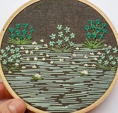 someone is holding up a small embroidery project with flowers and plants in the water on it