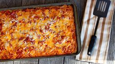 a square casserole dish with cheese and tomato sauce on top, next to a spatula