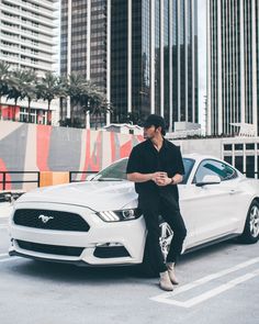 a man sitting on the hood of a white mustang car in front of tall buildings