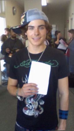 a young man in a black shirt and hat holding a white piece of paper with writing on it