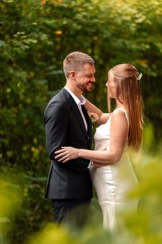 a man and woman standing next to each other in front of some green bushes with trees behind them