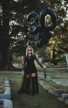 a woman standing in the grass holding some black balloons and wearing thigh high boots with her legs crossed