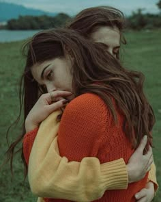 two young women embracing each other in a field with mountains in the background, one woman is wearing an orange and yellow sweater