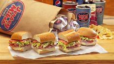 several sandwiches and chips on a wooden table with sodas in the backround