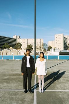 a man and woman standing in an empty parking lot with buildings in the back ground