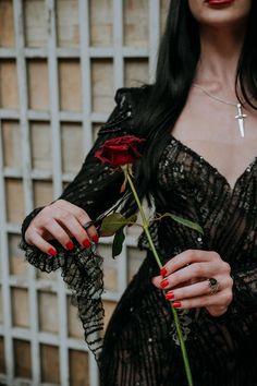 a woman with long black hair and red nails holds a rose in front of her face