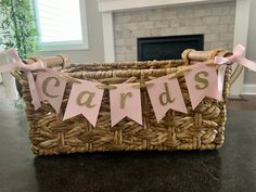 a wicker basket with pink and gold cards hanging from the handles, on a table in front of a fireplace