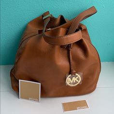 Michael By Michael Kors Large Grab Bag Is A Tan Bucket Bag Made Of Genuine Leather And Gold Hardware. The Circular Mk Logo In Gold Tag Is In Front Still Sealed In Plastic, Handles Are Both Joined In The Bag By A Band Of Golden Hardware (Pristine, No Scratches). Bag May Be Closed By A Golden Snap Closure. Bottom Measures Approximately 13” X 7”. Height Is 11 1/2 Inches. Bag Drop Is 10”. No Dust Bag. Nwt Caramel Travel Bag With Gold-tone Hardware, Small Backpack Purse, Gold Tag, Michael Kors Tote Bags, Black Leather Tote Bag, Mk Logo, In The Bag, Michael Kors Wallet, Grab Bag