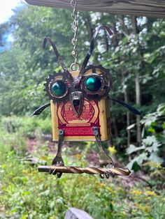 an owl shaped bird feeder hanging from a chain in front of some trees and bushes