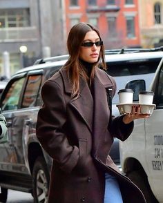 a woman holding two cups of coffee while walking down the street