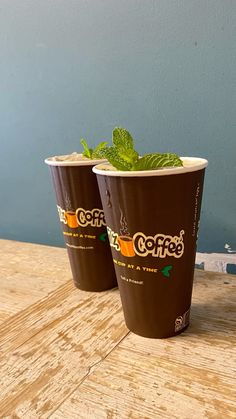 two coffee cups sitting on top of a wooden table next to a cup filled with green leaves