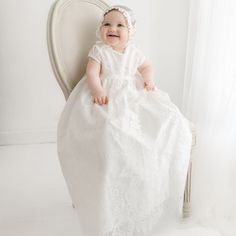 A smiling baby wearing a Victoria Puff Sleeve Christening Gown & Bonnet sits on a vintage, light-colored chair. The gown features ivory embroidered lace over a silk Dupioni lining. The The background is white and softly lit. Lace Bonnet, Baptism Gown, Ivory Silk, Christening Gowns, Silk Material, Pink Champagne, Scalloped Lace, Gowns With Sleeves, Fall Skirts