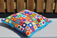 a multicolored crocheted pillow sitting on top of a white table next to a wooden fence