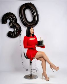 a woman in a red dress is sitting on a chair with a cake and balloons