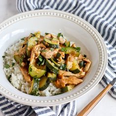 a white bowl filled with chicken, zucchini and rice next to chopsticks