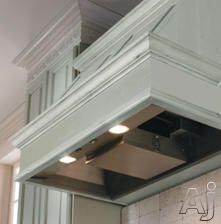 a kitchen with green cabinets and an overhead range hood over the stove top, in front of a brick wall