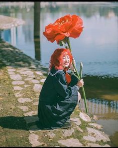 a woman with red hair sitting on the ground next to water and holding a flower