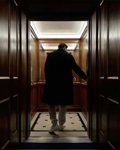 a man in a black coat and white pants is walking through an open wooden door