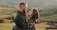 a man and woman standing close to each other in an open field with mountains in the background