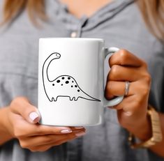 a woman holding a coffee mug with a drawing of a dinosaur on it's side