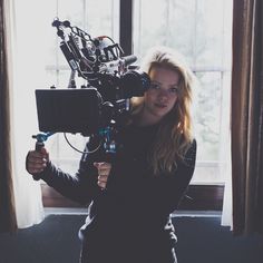 a woman standing in front of a window with a camera on her shoulder and holding a video camera