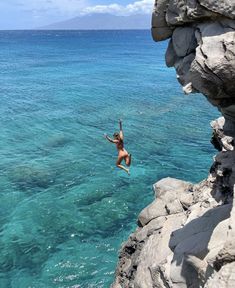 a person jumping into the ocean from a cliff near some rocks and blue water with mountains in the background