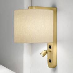 a wall light with a white shade on it's side and a bed in the background