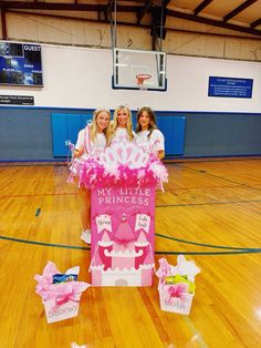 three girls in pink cheerleader outfits standing behind a cardboard sign on a basketball court