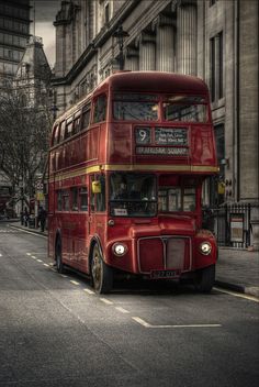 a red double decker bus driving down a street next to tall buildings on either side