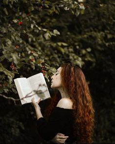 a woman with long red hair is reading a book in the dark, surrounded by trees