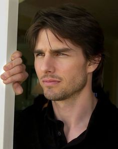 a man with long hair and black shirt leaning against a window sill looking at the camera