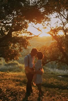 a man and woman standing in front of trees at sunset with the sun setting behind them