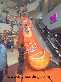 two children playing on an orange slide in a shopping mall with people walking around it