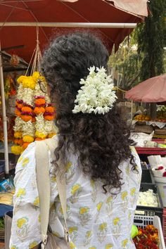 a woman standing in front of an open air market with flowers on her head and purse over her shoulder